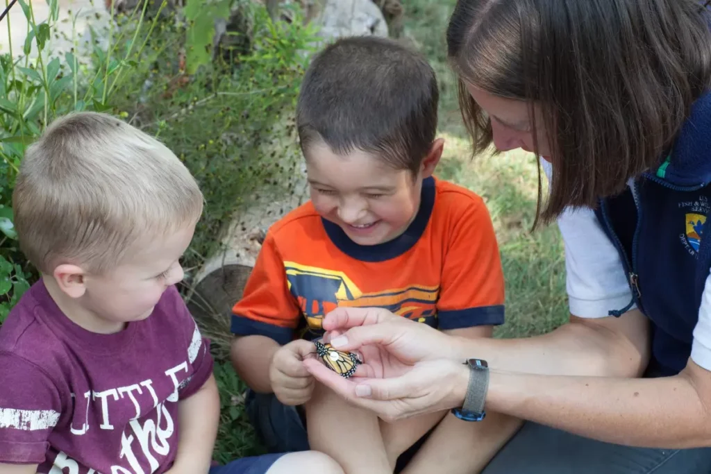 Two young children smiling while observing a recently tagged monarch butterfly being held by a woman.
